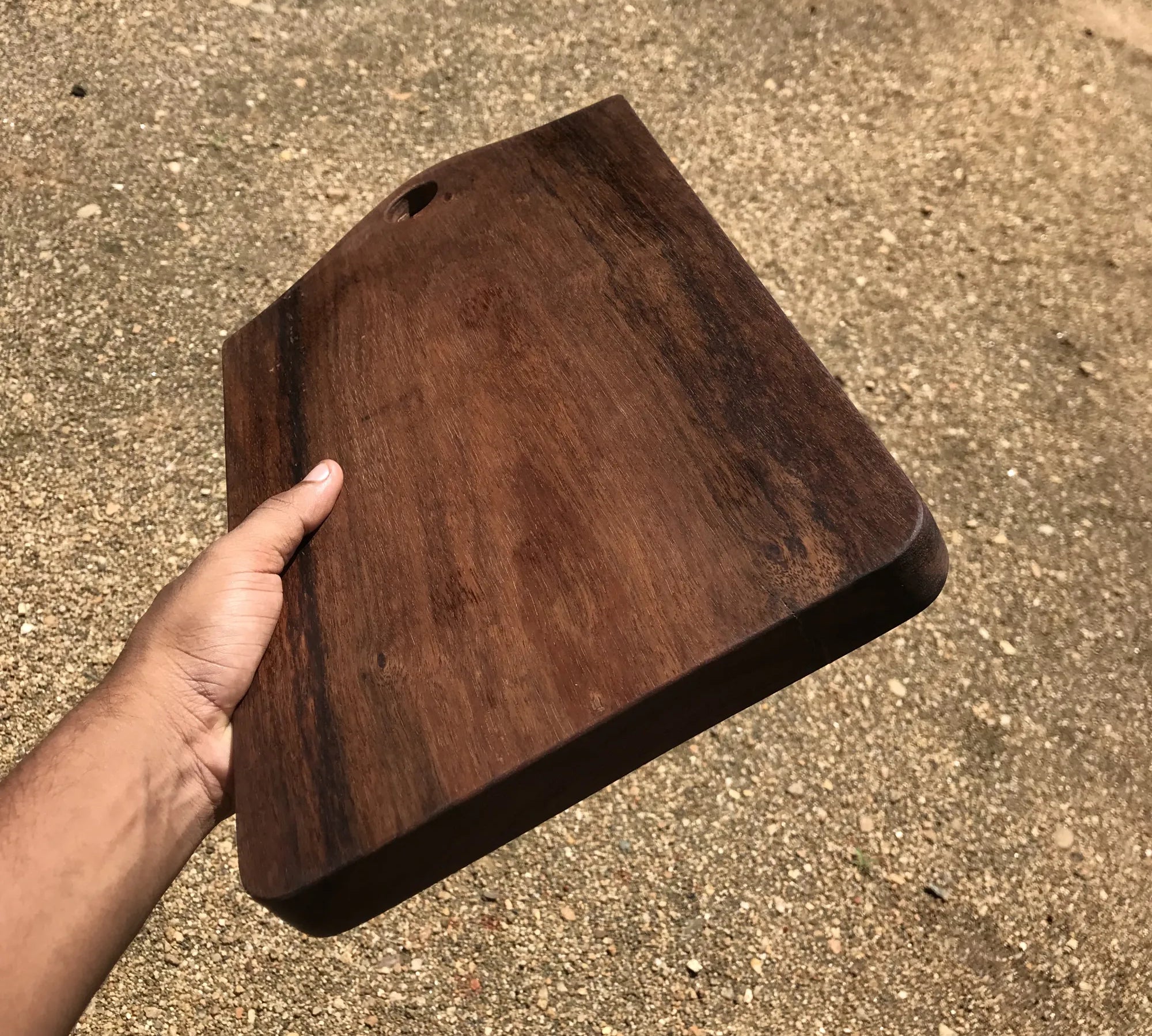 Hand holding a dark brown wood cutting board with rounded edges and a hole for hanging